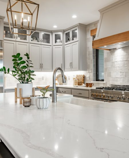 Beautiful home painted white with a modern farmhouse vibe. Large kithchen with white countertops, grey cabinets, stainless appliances, large white porcelain double sink farmhouse style. Lovely lighting fixtures, chandeliers, dining table with chairs. The white granite/marble counter has bar seating stools. There is a multiple burner gas stove with a large range hood fan, kitchen and dining room look toward the living room.